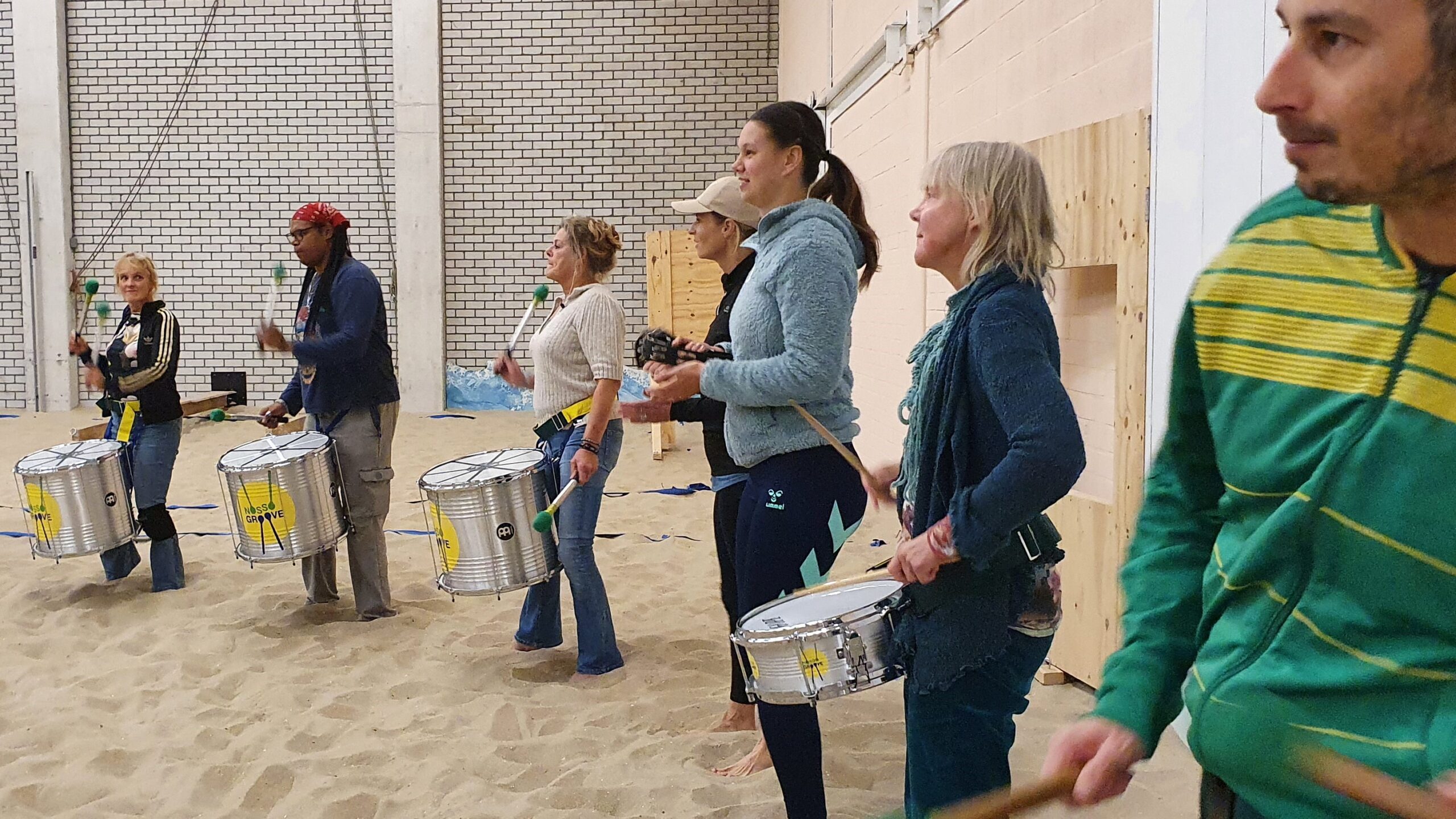 Zes personen met trommels op het zand tijdens de percussie workshop.
