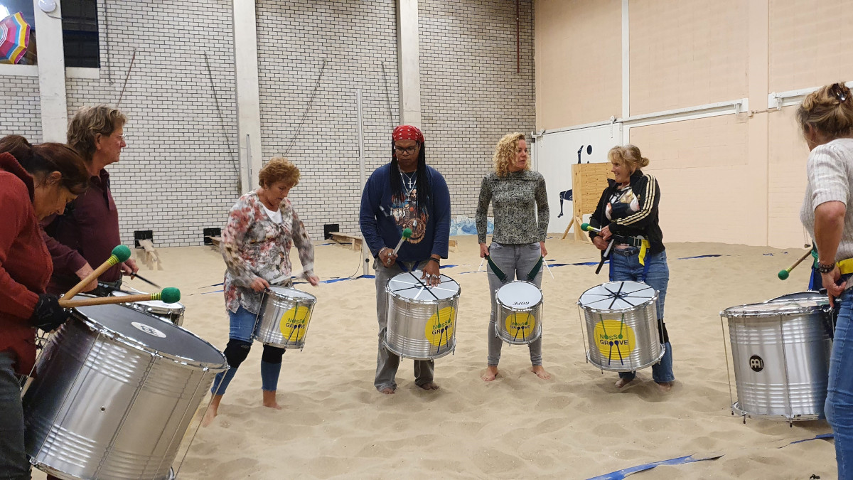 Acht personen die klaarstaan met hun trommels op het zand voor de percussie workshop.