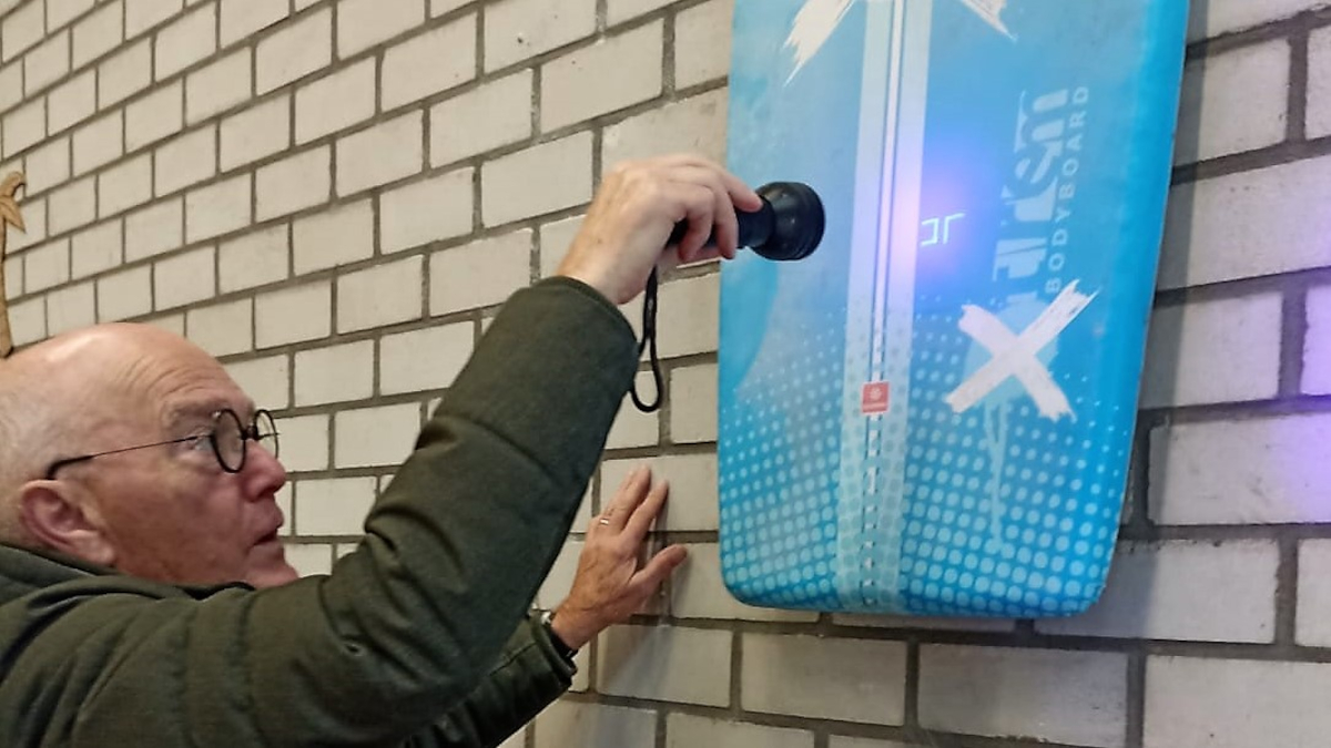 Een man schijnt met de blacklight zaklamp op het bodyboard op zoek naar aanwijzingen van het MasterMinds spel.