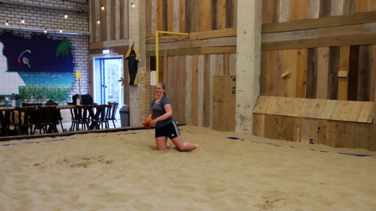 Een dame op haar knieën in het zand met een dodgeball in haar handen.