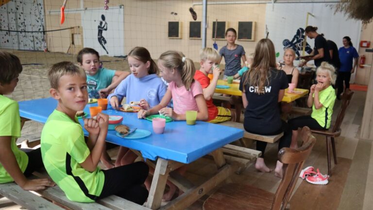 De kinderen van het zomerkamp eten lunch aan tafel.