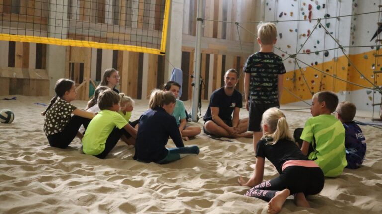 De kinderen zitten samen met de instructeur in een cirkel op het zand.