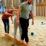 Teamuitje ideeën - Expeditie Robinson. Drie mannen balanceren op een balk terwijl ze worden bekogeld met ballen om ze er af te gooien tijden het teamuitje van Expeditie Robinson.