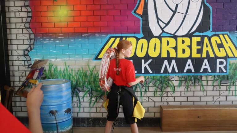 Aventuras Zomerkamp bij Indoor Beach Alkmaar. Een meisje staat in een surfpak bij het logo van Indoor Beach Alkmaar.